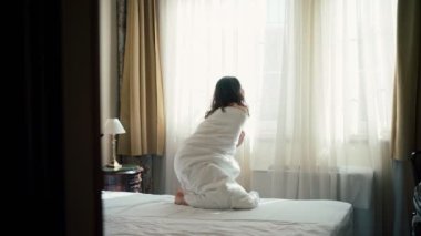 Battaniyeye sarılı genç bir kadın otel odasındaki pencerenin perdesini açar ve güneşli bir günde şehir manzarasının tadını çıkarır.