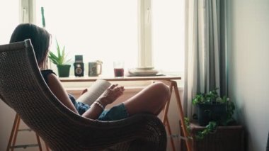 Pencerenin karşısındaki bir odada rahat bir hasır sandalyede oturan ve kitap okuyan bir kadının el kamerası görüntüsü.