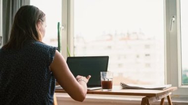 Pencerenin karşısındaki masada hasır bir sandalyede oturan bir kadının dizüstü bilgisayarda çalışırken çekilmiş el kamerasıyla çekilmiş bir fotoğrafı.