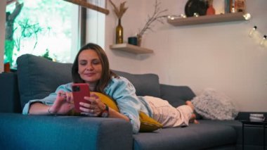 Oturma odasındaki kanepede uzanmış akıllı telefonunu kullanan genç bir kadın.