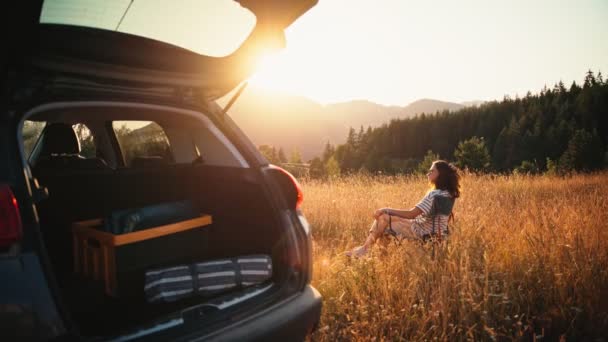 折りたたみ椅子に座っている若い女性の映画撮影と彼女のソロカー旅行中に夕日を楽しむ — ストック動画