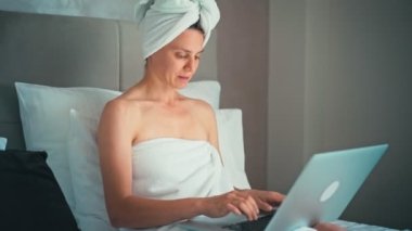 Kafasında havlu olan genç bir kadın iş gezisi sırasında otel odasında dizüstü bilgisayarla çalışıyor.