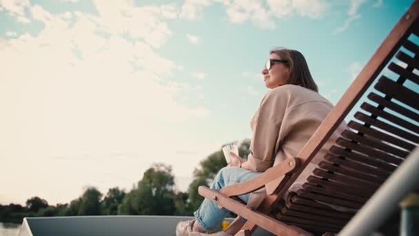 一位戴着太阳镜的年轻的高加索女人坐在游艇顶层的木制日光浴台上欣赏美景 — 图库视频影像