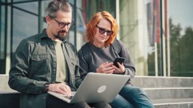 Bilgisayarlarını ve akıllı telefonlarını kullanan iki beyaz erkek ve kadın arkadaş modern iş binasının merdivenlerinde otururken projeyi tartışıyorlar..