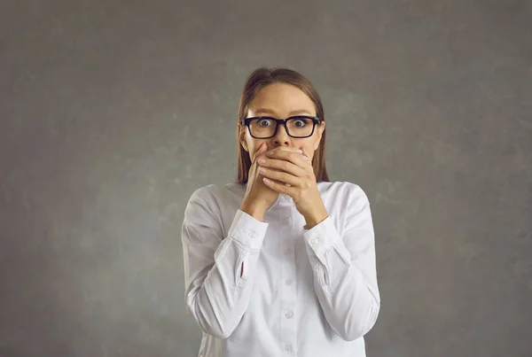 Young shocked woman in eyeglasses with hand over mouth studio portrait shot — Stock Photo, Image