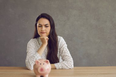 Bir kadın domuz kumbarası olan bir masada oturuyor ve parasını nereye harcayacağını planlıyor.