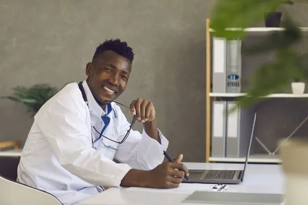 Junger schwarzer Arzt sitzt mit Laptop am Schreibtisch, blickt in die Kamera und lächelt — Stockfoto