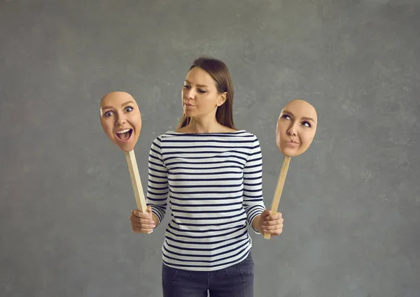 Woman stands on a gray background and holds two faces with different emotions and moods. — Stock Photo, Image