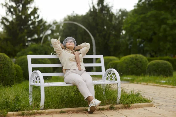 木製のベンチに座っている幸せなリラックスしたシニア女性は公園で晴れた日を楽しむ — ストック写真