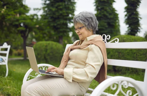 Alte Dame benutzt Laptop und ruht sich im Park aus, während sie auf Bank sitzt — Stockfoto