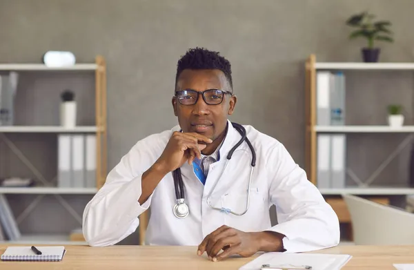 Porträt eines jungen schwarzen Arztes mit Brille, der am Schreibtisch sitzt und dem Patienten zuhört — Stockfoto