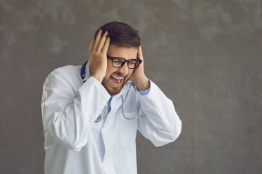 Kızgın doktor bağırıyor ve kulaklarını kapatıyor. Gri arka planda elleri kapalı.