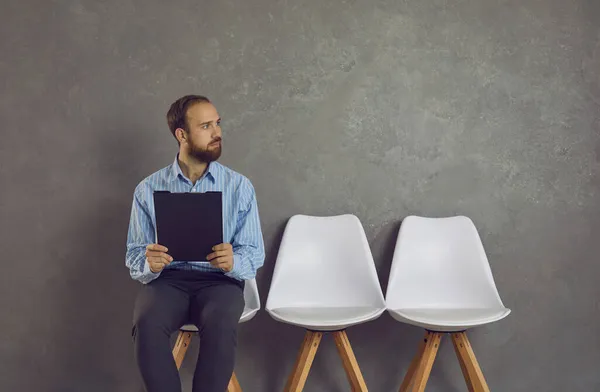 Mladý muž sedí na židli, drží životopis a dívá se jinam a čeká na pracovní pohovor — Stock fotografie