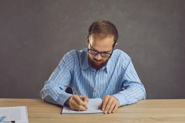 Ernstige jongeman met een bril zittend aan een tafel en aantekeningen makend in zijn notitieboekje — Stockfoto