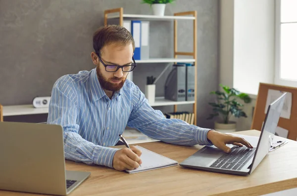 Ernster, konzentrierter Mann sitzt mit Laptop am Schreibtisch und macht sich Notizen in Notizbuch — Stockfoto