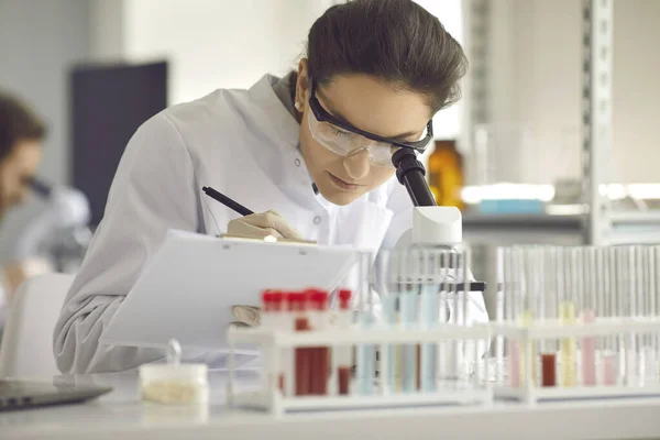 Close-up vrouw wetenschapper werken in laboratorium met microscoop en klembord — Stockfoto