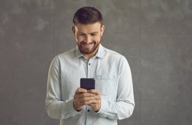 Gri arkaplanda duran akıllı telefon ekranına bakan tatmin olmuş telefon kullanıcısı