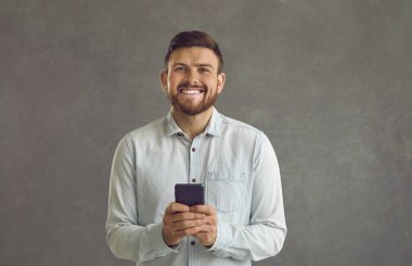 Akıllı telefon ve gülümseme kullanan mutlu genç adam gri arka planda tek başına duruyor.