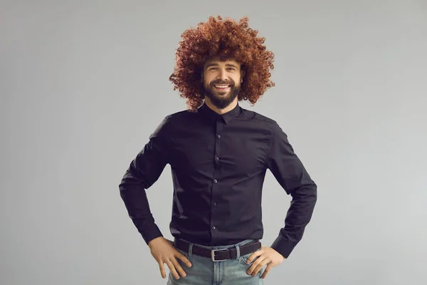 Funny happy man wearing curly brown wig standing hands oh hips on gray background — Stock Photo, Image