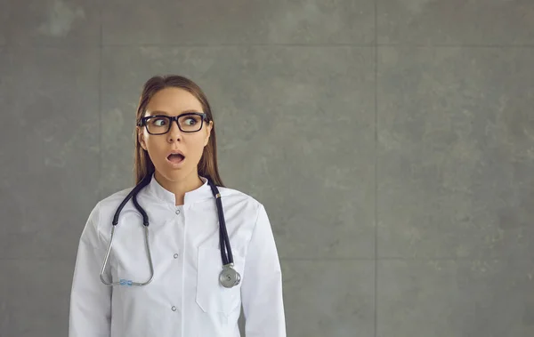 Шокированная молодая женщина-врач с открытым ртом смотрит в сторону студийной фотографии — стоковое фото