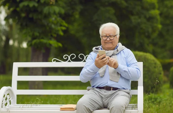 Gelukkig senior man met behulp van een mobiele telefoon tijdens het zitten op een bank in een groene zomer park — Stockfoto