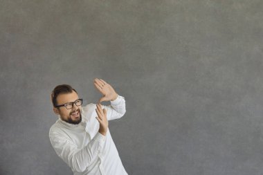 Komik gülümseyen, korkmuş adam yüzünü kapayarak gri arka planda duruyor.