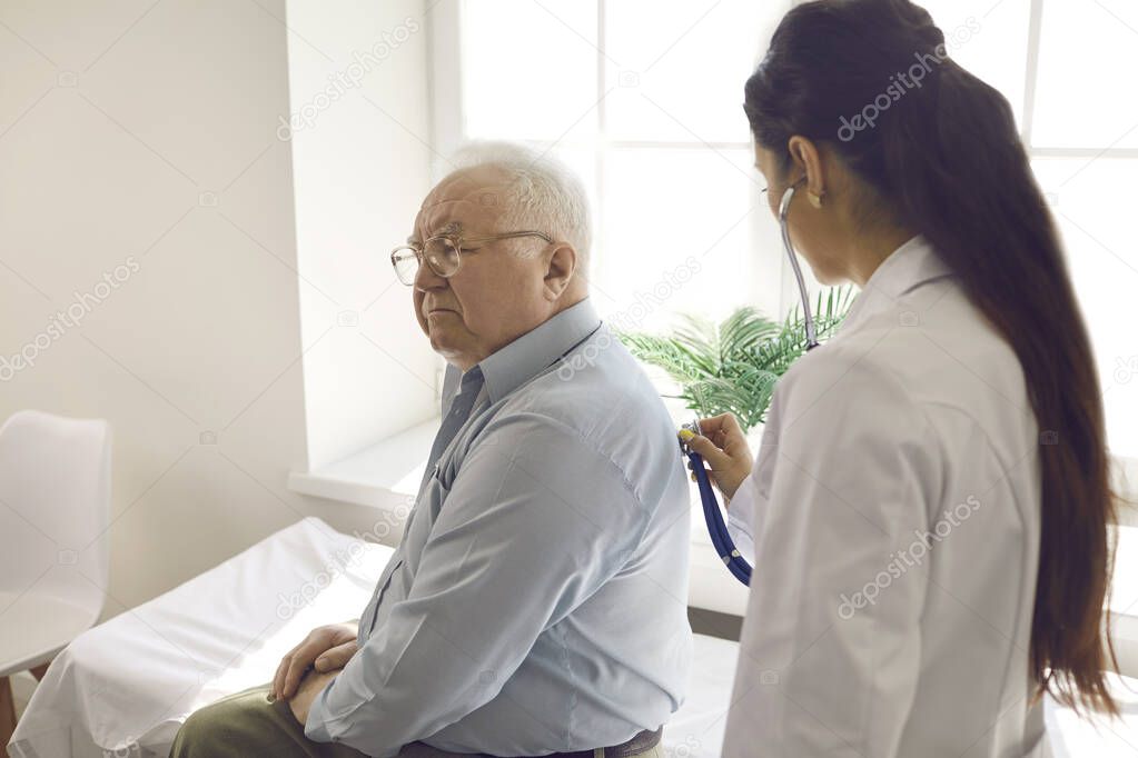 Female doctor examining elderly male patient by stethoscope at hospital ward