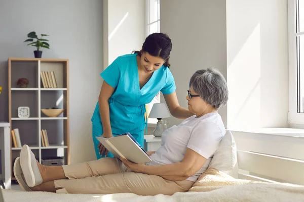 Infirmière en soins à domicile parlant à une aînée détendue assise sur son lit et lisant un livre — Photo