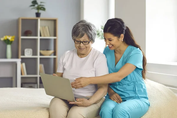 Amichevole infermiera di assistenza domiciliare insegnare felice donna anziana di utilizzare il computer portatile moderno — Foto Stock