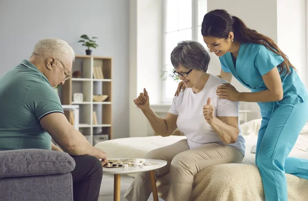 Aide soignante regarder couple de ses patients âgés heureux jouer dames — Photo