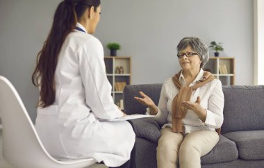 Yaşlı kadın hasta, evde kanepede oturan hemşire hemşireyle konuşuyor.