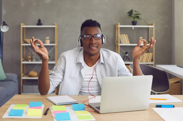 Un Afro-Américain détendu écoute de la musique sur un casque et médite pendant qu'il est assis au travail. — Photo