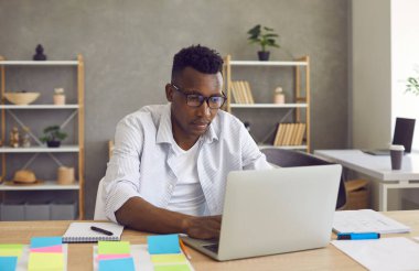 Afro-Amerikalı yönetici laptopta yazı yazıyor. Ofis masasında oturuyor.