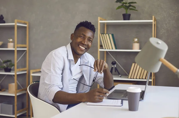 Homme d'affaires noir souriant regardant la caméra assis au bureau avec ordinateur portable dans le bureau à la maison — Photo