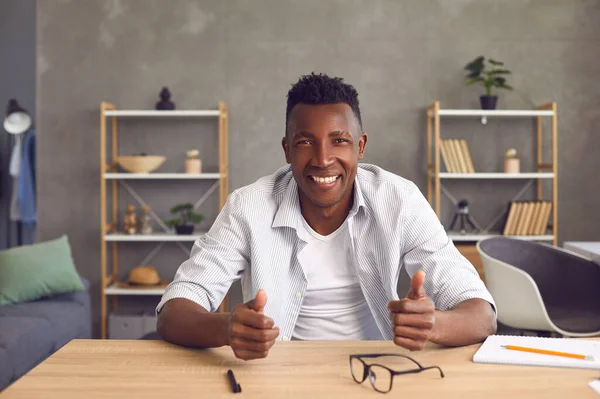 Heureux jeune homme africain amical assis à la table dans le bureau à la maison et souriant à la caméra — Photo