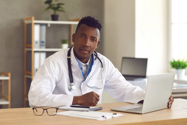 Glücklicher schwarzer Arzt sitzt am Schreibtisch und nutzt Laptop für Videoanruf mit Patient — Stockfoto