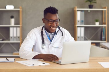 Ofis masasında oturan mutlu siyah doktor hastayla görüntülü konuşma için dizüstü bilgisayar kullanıyor.