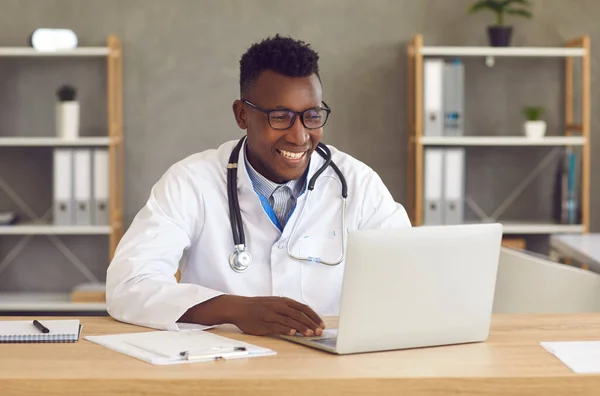 Glücklicher schwarzer Arzt sitzt am Schreibtisch und nutzt Laptop für Videoanruf mit Patient — Stockfoto