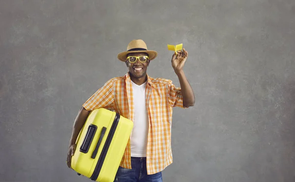 Studio portret van gelukkige zwarte man met papieren vliegtuig en gele reiskoffer — Stockfoto