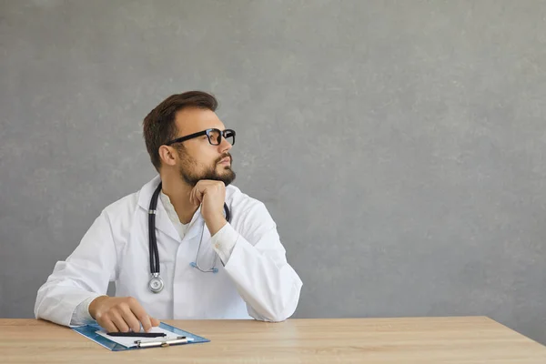 Sonhador médico masculino pensativo em vestido médico branco terno sentar na mesa olhar para o lado — Fotografia de Stock