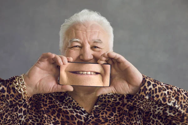 Uomo anziano divertente che copre il viso con il telefono cellulare con immagine ingrandita di sorriso perfetto — Foto Stock