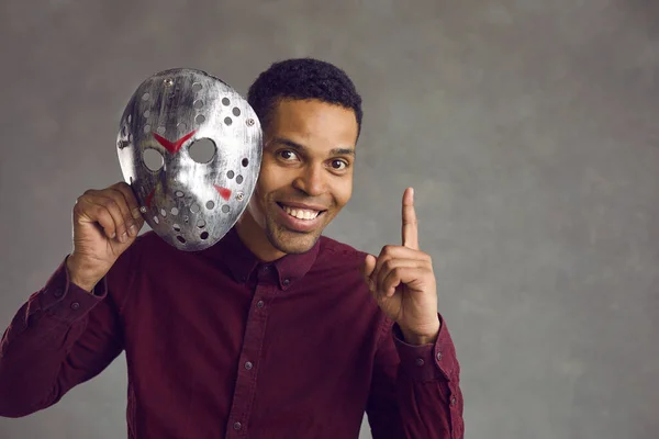 Happy handsome young black man takes off his ugly scary mask, points finger up and smiles — Stock Photo, Image