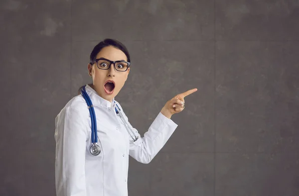 Шокированный доктор в белом халате и очках указывает пальцем на копировальный фон — стоковое фото