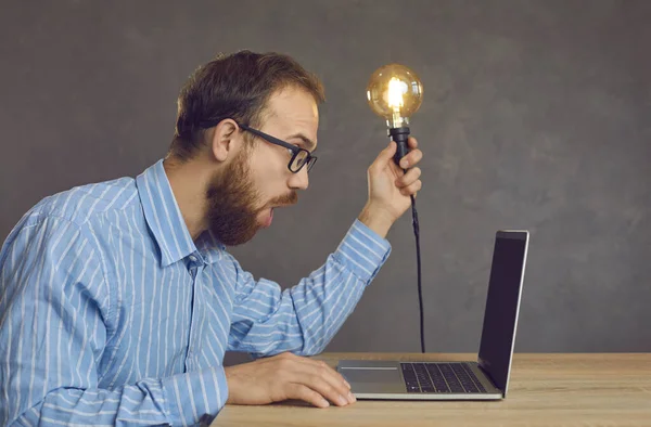 Boční pohled na legračního překvapeného muže sedícího před laptopem se žárovkou v rukou. — Stock fotografie