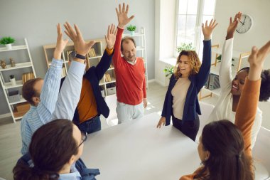 Farklı etnik çeşitlilikte farklı yaş grupları masanın etrafında el kaldırarak duruyorlar.