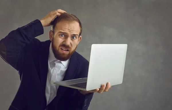Kafası karışmış bir iş adamı, elinde bir dizüstü bilgisayarla gri bir arka planda dikilirken kafasını kaşıyor.. — Stok fotoğraf