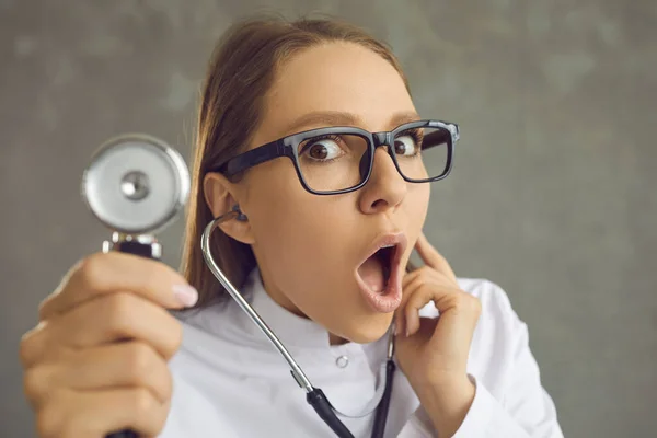 Шокированная женщина-врач с удивленным лицом поднимает стетоскоп, слушая сердцебиение. — стоковое фото