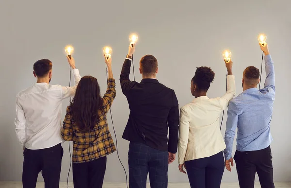 Vista posterior de un grupo de jóvenes empresarios diversos que sostienen bombillas brillantes —  Fotos de Stock