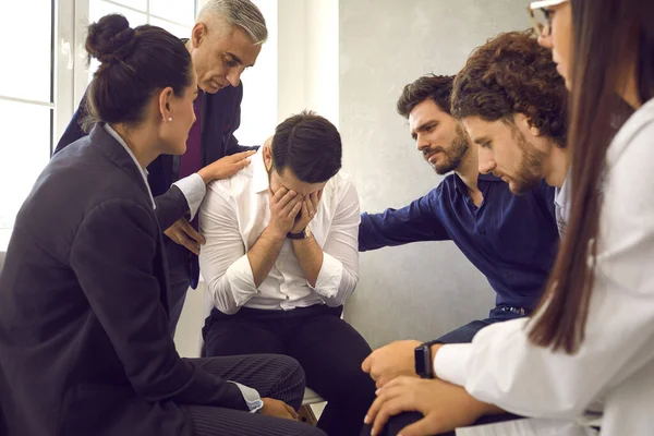 People in group therapy supporting distraught crying man, comforting him, helping him deal with grief and problems. Young man hides face and cries while telling his story sitting among other patients