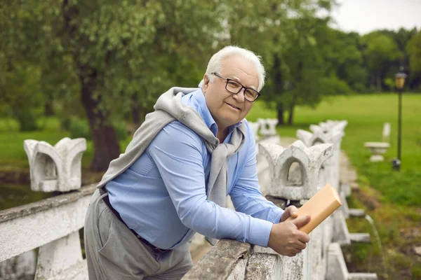 Portret van een positief lachende oudere man die in een zomerpark loopt met een boek in zijn handen. — Stockfoto